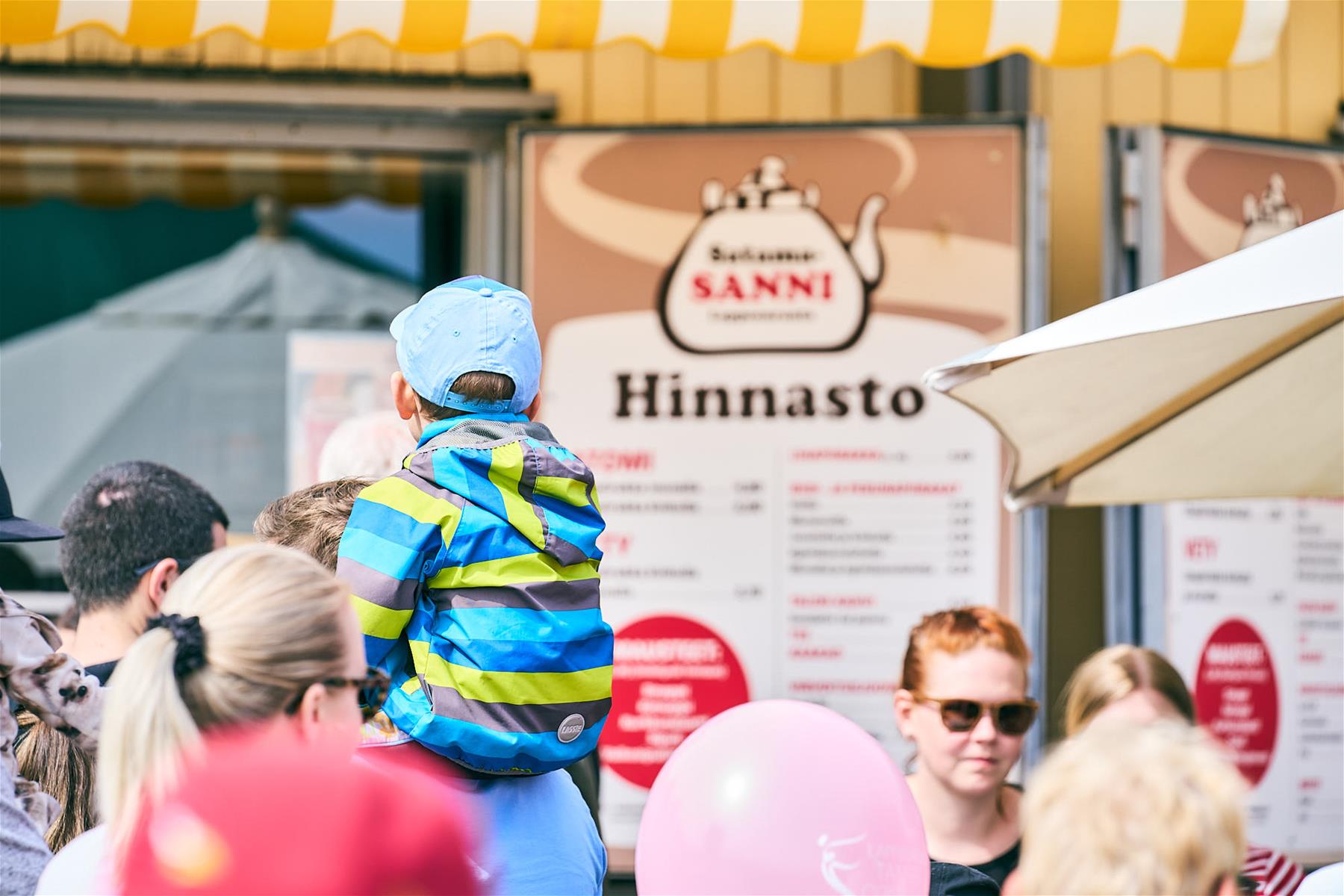Ihmisiä keltaisen vetykojun edessä. Jonottamassa aikuinen, jonka hartioilla istuu lapsi, jolla on raidallinen takki.  