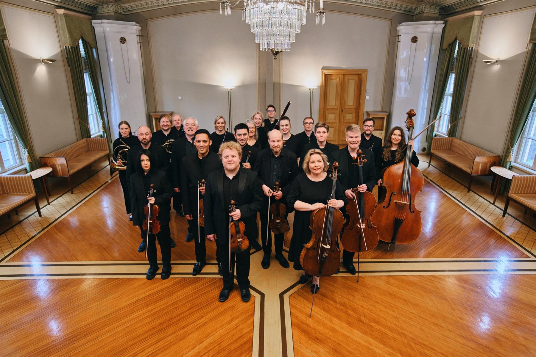 Lappeenrannan kaupunginorkesterin muusikoiden ryhmäkuva. Muusikot seisovat juhlavassa salissa mustiin pukeutuneina ja soittimet kädessä.